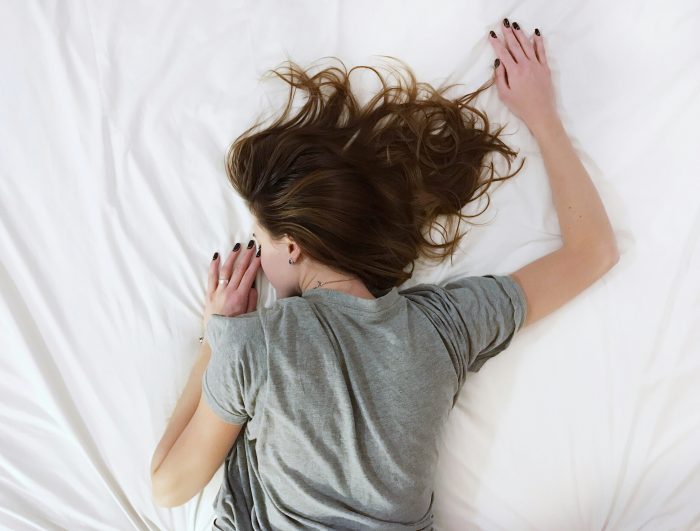 a woman laying face down on a bed, she has mousey brown hair and her nails are painted black, she is wearing a grey t-shirt. 